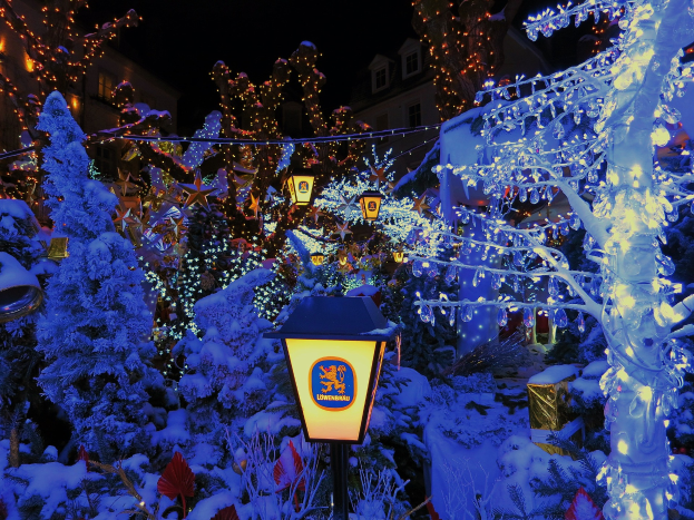 Ein Weihnachtsbaum steht in der Mitte einer verschneiten Straße bei Nacht, beleuchtet von Lichtern und umgeben von schneebedeckten Pflanzen, mit Gebäuden im Hintergrund, die beleuchtete Fenster aufweisen.