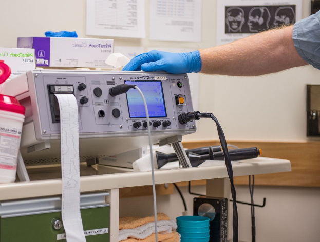 Eine Person in blauen Handschuhen arbeitet an einem medizinischen Gerät auf einem Tisch, der mit verschiedenen Gegenständen übersät ist, mit Papieren an der Wand im Hintergrund, was auf eine Krankenhausumgebung hinweist.