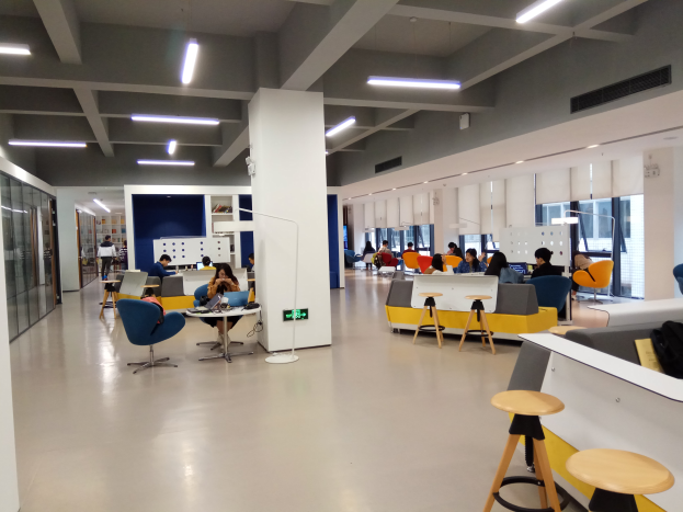 Ein geräumiges Großraumbüro mit Menschen an Tischen und Stühlen sitzend, Laptops und Gegenständen auf den Tischen, einer Theke mit einer Tasche und anderen Gegenständen auf der rechten Seite und Glaswänden mit Deckenleuchten im Hintergrund.