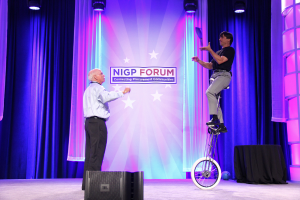 Eine Person steht auf der Bühne und spricht, während eine andere Person auf einem Fahrzeug hinter ihnen steht, unter blauen Vorhängen, Scheinwerfern und einem Banner.