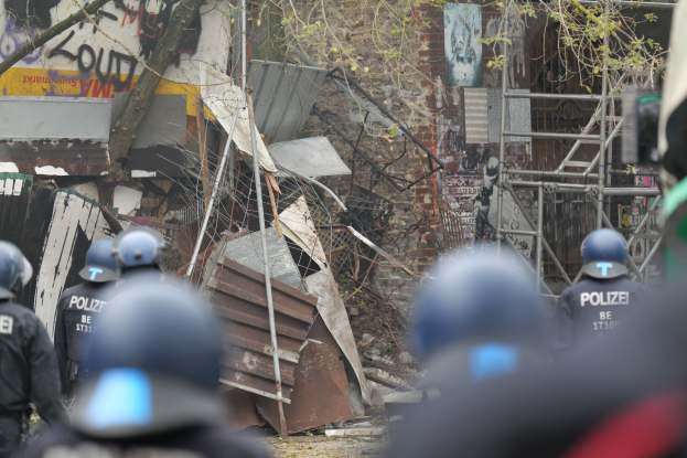 Eine Gruppe von Polizisten mit Helmen steht vor einem teilweise abgeräumten Gebäude mit Text an seiner Wand, umgeben von Bäumen und Masten.