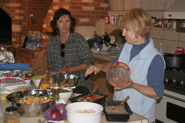 Zwei Frauen stehen in einer Küche umgeben von verschiedenen Lebensmitteln und Küchenutensilien auf der Arbeitsplatte, mit einem Gasherd auf der rechten Seite und Flaschen, einem Kamin und einer Backsteinwand im Hintergrund.