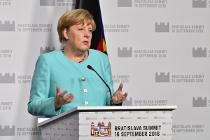 Bundeskanzlerin Angela Merkel hält eine Rede auf dem Bratislava-Gipfel, vor einem Podium mit zwei Mikrofonen stehend, mit einem Banner und einer Flagge im Hintergrund.