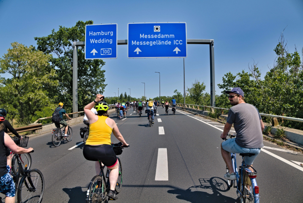 Radfahrer mit Helmen fahren auf einer Straße mit einem Geländer auf der linken Seite und Bäumen auf der rechten Seite unter einem klaren blauen Himmel mit Laternen im Hintergrund; ein Schild oben zeigt eine Radtour in Hamburg.