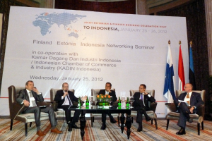 Eine Konferenzbühne mit Menschen, Stühlen, einem Mikrofon, Kabeln, einem Tisch, Flaschen, Blumen, Gläsern und anderen Gegenständen, mit Fahnen und einem Banner in der Mitte und einem Gebäude mit Vorhängen im Hintergrund.