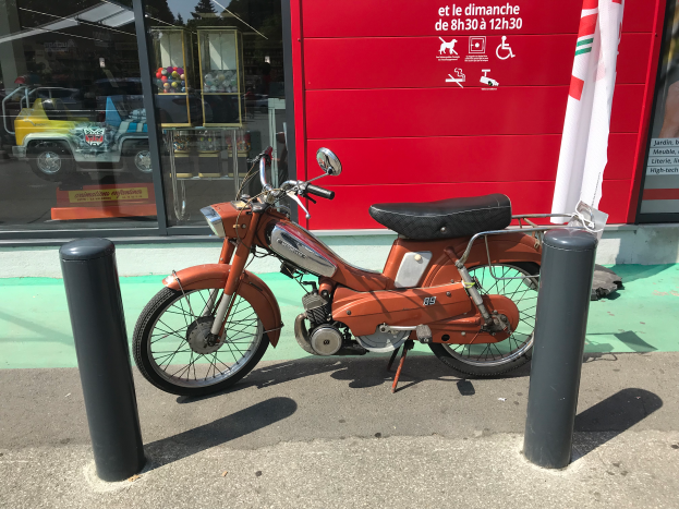 Ein oranger Moped steht auf der Straße vor einem roten Gebäude mit Glaswänden, mit zwei schwarzen Pfählen im Vordergrund und einer Flagge mit Text an der Wand rechts.