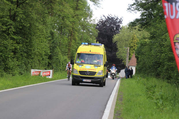 Ambulanz fährt auf einer Straße mit Fahrradfahrern nebenher, Gras und Bäume auf beiden Seiten, Häuser, Masten und einen klaren blauen Himmel im Hintergrund.