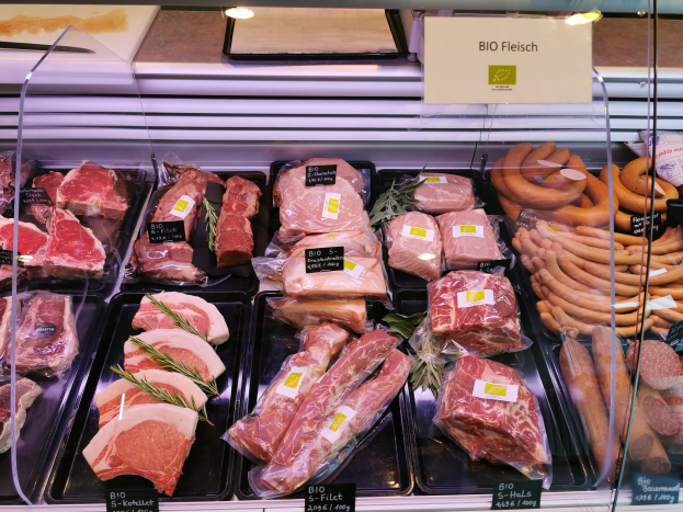 Ein Auslageglas voller verschiedener Fleisch- und Wurstwaren in Schalen, jeweils mit Preisschildern, in einem Metzgerladen mit einer Texttafel im Hintergrund.