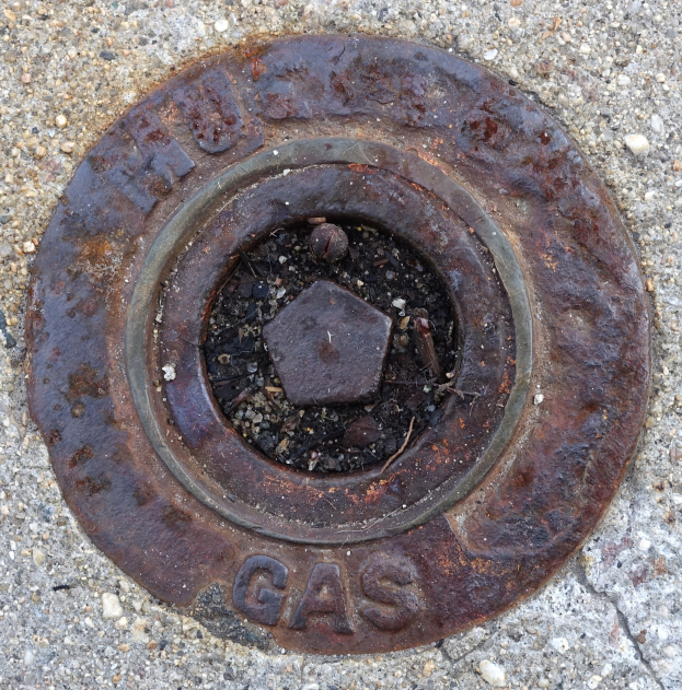 Ein verrosteter Metall-Schachtobdeckel auf dem Boden mit dem Wort 'Gas' darauf geprägt.