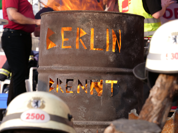 Eine Gruppe von Menschen um ein Feuerhydrant mit der Aufschrift "Berlin" herumstehend mit einem brennenden Fass im Vordergrund, Helme unten und ein hölzernes Objekt rechts.