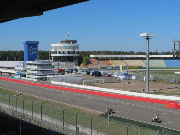 Gruppe von Motorradfahrern auf einer Rennstrecke, umgeben von einem Metallzaun, Gras und einem Stadion mit einem bewölkten Himmel.