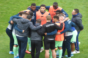 Eine Gruppe von Männern in Jacken steht im Kreis auf einem Fußballfeld.