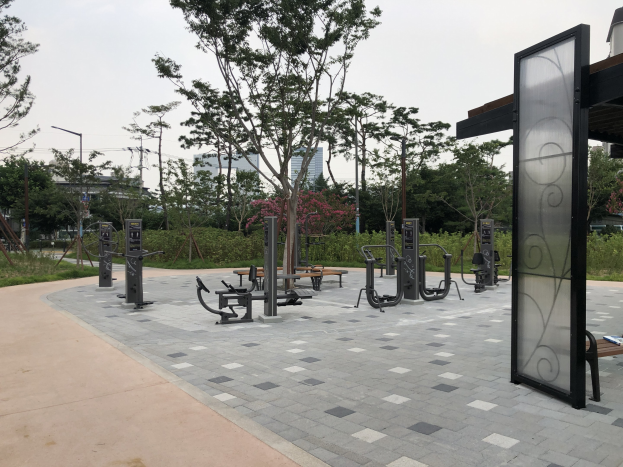Outdoor park featuring benches, trees, plants, grass, fitness equipment, poles, lights, wires, and buildings under a sky backdrop.
