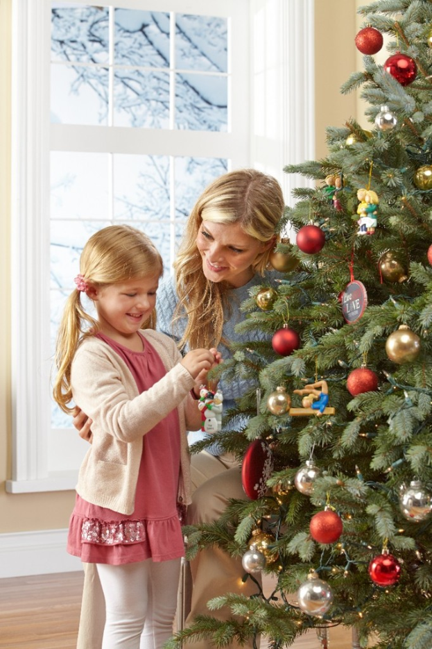 Ein geschmückter Weihnachtsbaum steht in einem Haus mit einer Frau und einem Kind im Vordergrund, einem Fenster und einer Wand im Hintergrund und Bäumen, dieoutside des Fensters sichtbar sind.
