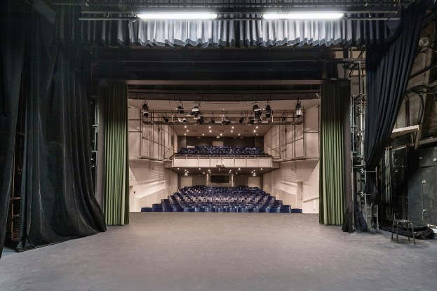 Ein großer Zuschauerraum mit schwarzen Vorhängen, einer Bühne, Deckenlampen und Stühlen, mit Gegenständen auf der rechten Seite.