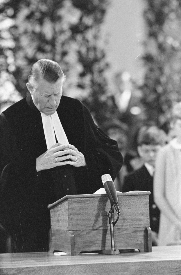 Ein Mann in schwarzer und weißer Robe steht an einem Podium mit einem Mikrofon, umgeben von ein paar Menschen und Bäumen im Hintergrund.