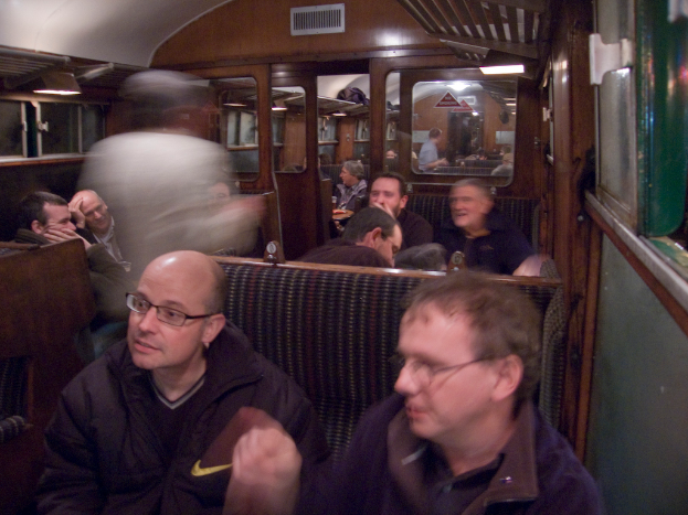 Zwei Männer sitzen auf einem Sitz in einem Zug, mit einer Gruppe von Menschen dahinter, einem Eingang zwischen Abteilen oben und einem Fenster rechts.