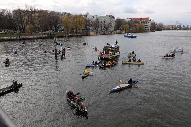 Eine Gruppe von Menschen in Kanus, die einen Fluss hinunterpaddeln, mit einem Geländer auf der linken Seite, Bäumen, Gebäuden, Fahrzeugen und Pfählen im Hintergrund unter einem klaren blauen Himmel.