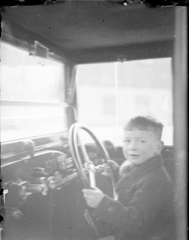 Ein junger Junge sitzt auf dem Fahrersitz eines Autos und hält das Lenkrad mit konzentrierter Miene in einer Schwarz-Weiß-Fotografie.