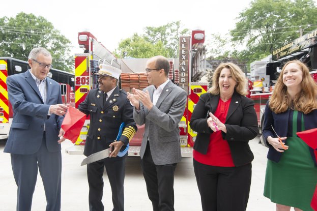 Gruppe von Menschen bei einer Ehrung für die Alexandria Fire Department, die vor einem Feuerwehrauto klatschen und lächeln, mit Bäumen und einem klaren Himmel im Hintergrund.