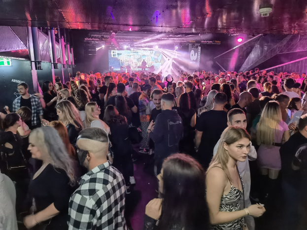 Eine große Gruppe von Menschen in einer Diskothek mit Deckenleuchten und Säulen, mit einem Bildschirm im Hintergrund, der als Absolut Nightlife-Event gekennzeichnet ist.