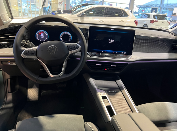 Das Innere eines Volkswagen e-Golf, mit einem Lenkrad, Armaturenbrett, Bildschirmen, Knöpfen, Sitzen und Glastüren, mit einem Blick auf andere Autos, Gebäude, Bäume, Pfosten und den Himmel durch die Glastüren.