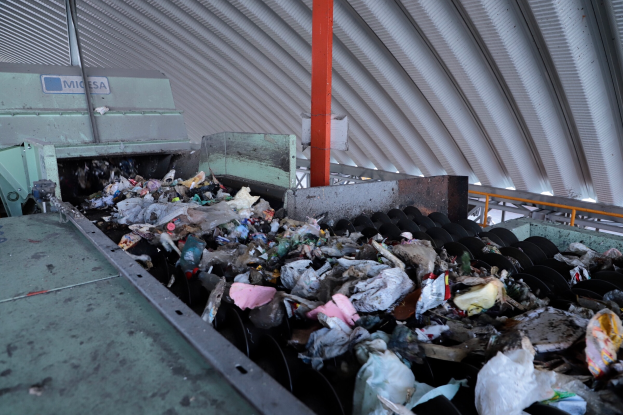 Ein großer Müllhaufen auf einem Förderband in einer Recyclinganlage, mit einer Maschine im Vordergrund und einem Metalldach darüber.