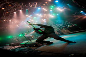 Ein musikalisches Konzert mit drei Performern auf der Bühne: zwei Gitarristen rechts mit Mikrofonen davor, ein Hintergrund mit "MOLLEY" und zwei Schlangenlogos sowie Scheinwerfer darüber.