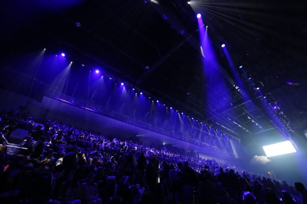 Eine große Menschenmenge sitzt vor einer Bühne mit Deckenbeleuchtung, mit einem Bildschirm auf der rechten Seite.