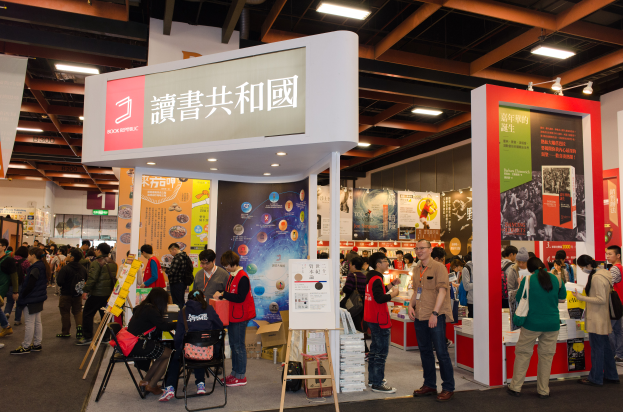 Eine große Gruppe von Menschen an einem Messestand mit Stühlen, Brettern, Bannern und Deckenleuchten, die 'Book Republic Hong Kong Book Fair' zeigt.