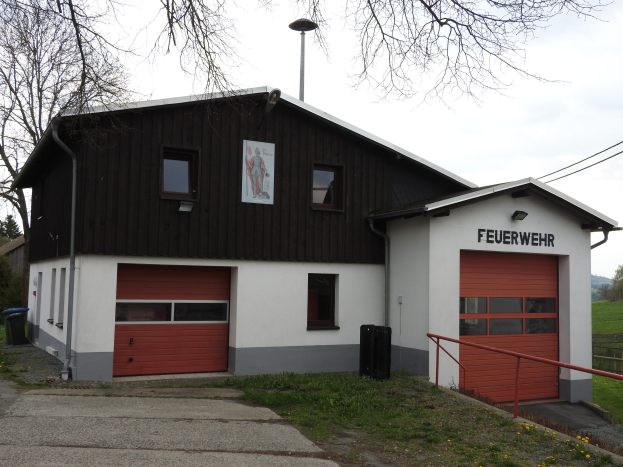 Rote und weiße Feuerwache-Gebäude mit einer Straße, Gras, gelben Blumen und Bäumen im Vordergrund und dem Himmel im Hintergrund.