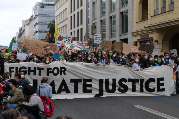 Eine große Gruppe von Menschen marschiert durch eine Straße und hält ein Transparent mit der Aufschrift "Kampf für Hassjustiz", einige tragen Masken und halten Schilder, und im Hintergrund sind Gebäude, Bäume und ein klarer blauer Himmel zu sehen.