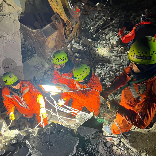 Gruppe von Rettungskräften in orangefarbenen Uniformen und grünen Helmen beseitigt Trömer von einem eingestürzten Gebäude mit einem Kran im Hintergrund.