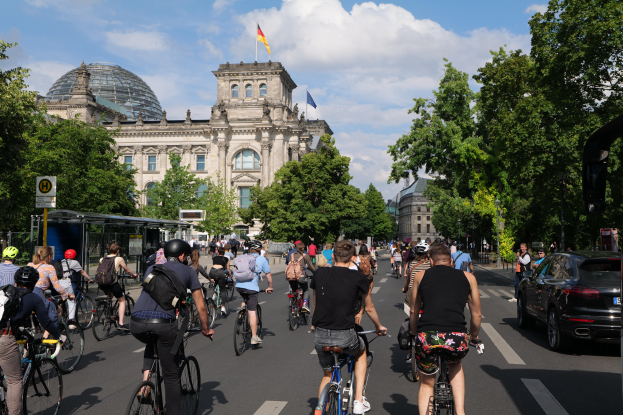 Eine Gruppe von Menschen, die Fahrräder auf einer von Bäumen gesäumten Straße in Berlin, Deutschland, fährt, mit Gebäuden, einer Bushaltestelle auf der rechten Seite und einer Flagge, die auf einem der Gebäude weht, unter einem bewölkten Himmel.