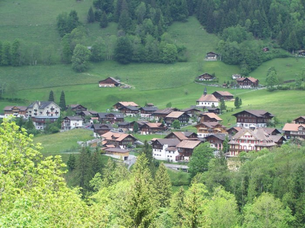 Kleines Dorf mit bunten Häusern in einer grünen Talsenke umgeben von Bäumen und Gras.
