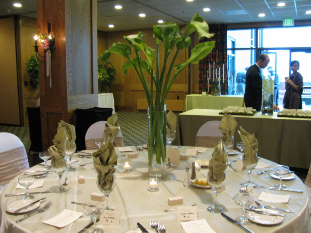 Ein Tisch mit weißer Decke, Tellern, Besteck und einem Glas, umgeben von Stühlen, mit einem grünen Tisch dahinter; ein Mann in einem schwarzen Blazer und eine Frau in der Nähe einer Tür mit der Aufschrift 'Ausgang', mit zwei Männern dahinter.