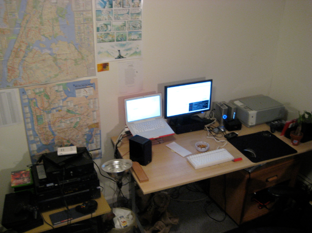 Ein Raum mit einem Tisch, auf dem ein Laptop, ein Lautsprecher, ein Monitor, eine Tastatur, ein Ball, eine Maus, ein Scanner und Papiere liegen; ein weiterer Tisch mit Elektronikgeräten in der linken unteren Ecke; und eine Wand mit Charts und Postern im Hintergrund.