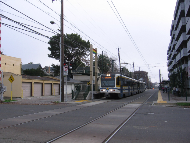 Straßenbahn fährt durch eine städtische Straße mit hohen Gebäuden, Strommasten, Fußgängern, Bäumen und einem klaren blauen Himmel.