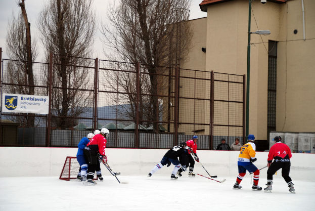 Menschen, die auf einem Eisstadion Hockey spielen, mit Gebäuden, Bäumen, einer Straßenlaterne, einem Namensschild und Zäunen im Hintergrund unter dem Himmel.