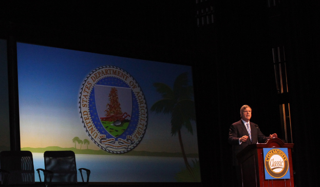 Ein Mann in einem weißen Hemd und schwarzem Blazer steht an einem Rednerpult, möglicherweise sprechend, mit Stühlen im Vordergrund und einem schwarzen Hintergrund, der möglicherweise eine Tafel oder ein Projektionsschirm ist, was auf eine Konzertsitzung hindeutet.