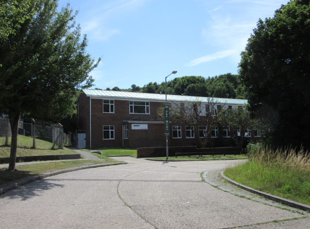 Ein Schulgebäude mit Fenstern befindet sich in der Mitte eines Parks, umgeben von saftigem Grün, mit einem Laternenmast, einem Schild, einer Baumgruppe, einem Zaun und einem bewölkten Himmel im Hintergrund.
