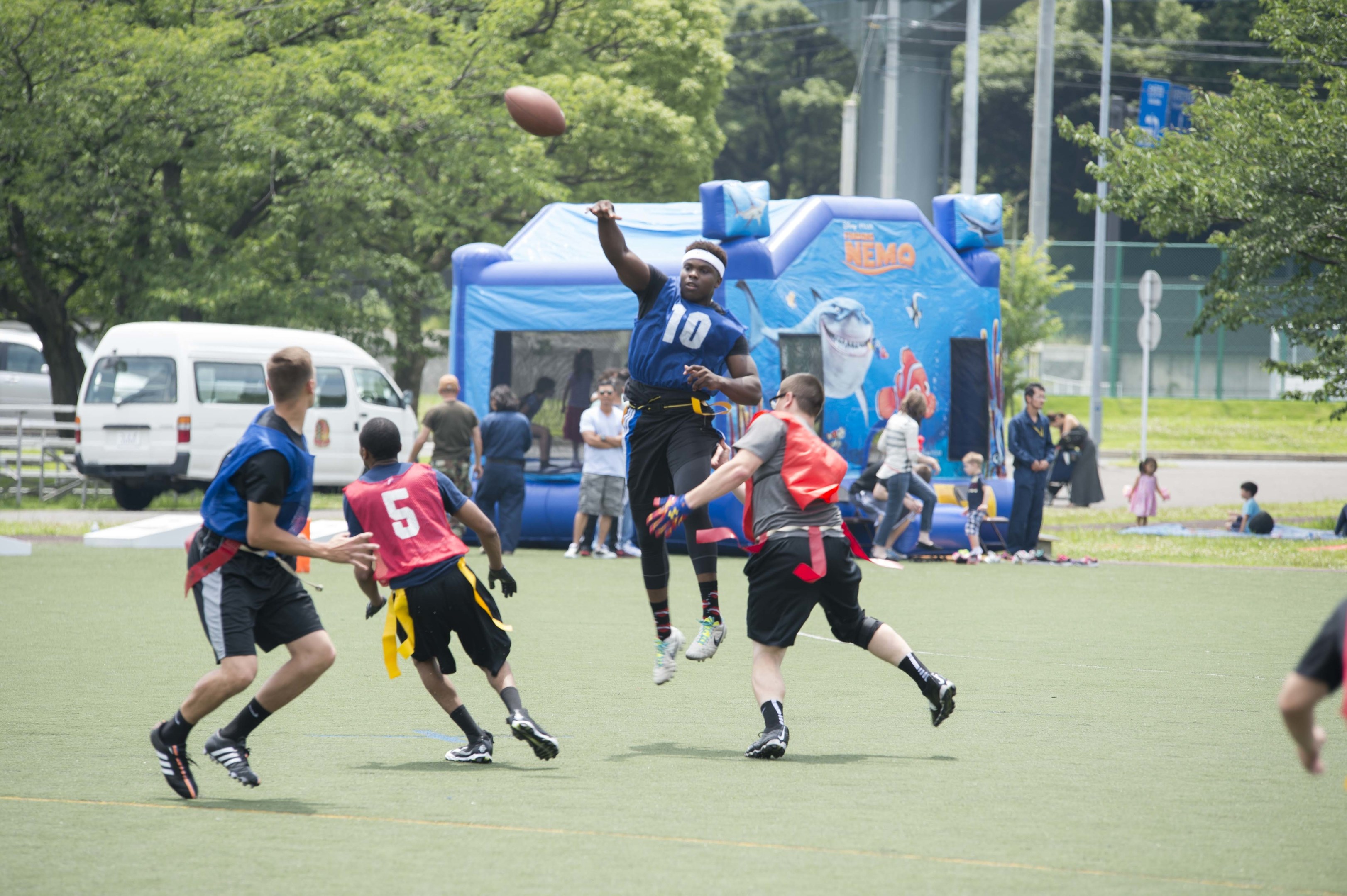 Gruppe junger Männer beim Fußballspielen auf einem Rasenfeld mit Zuschauern, einem Zelt, Fahrzeugen, Bäumen und einem Zaun im Hintergrund vor einem sichtbaren Himmel.