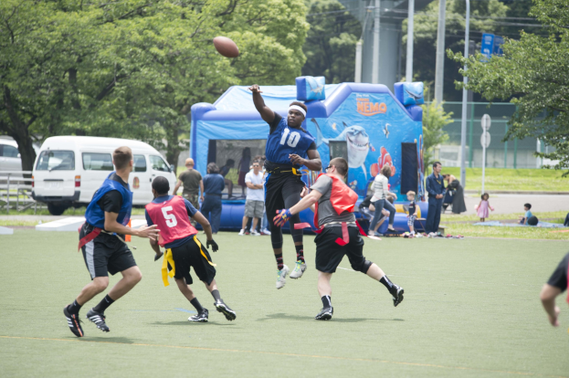 Gruppe junger Männer beim Fußballspielen auf einem Rasenfeld mit Zuschauern, einem Zelt, Fahrzeugen, Bäumen und einem Zaun im Hintergrund vor einem sichtbaren Himmel.