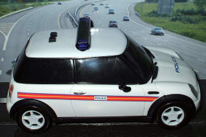 Ein weißes Polizeiauto ist im Vordergrund geparkt, mit mehreren Autos, die sich auf der Straße dahinter bewegen, umgeben von Gras und Bäumen auf beiden Seiten, in einer ländlichen Umgebung.