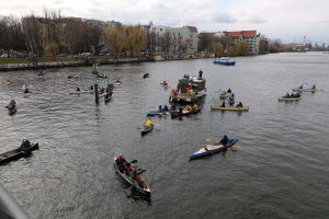 Eine Gruppe von Menschen in Kanus, die einen Fluss hinunterpaddeln, mit einem Geländer auf der linken Seite, Bäumen, Gebäuden, Fahrzeugen und Pfählen im Hintergrund unter einem klaren blauen Himmel.