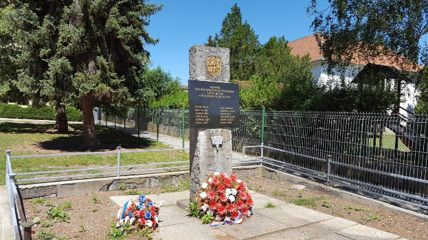 Ein Denkmal für die Opfer der Tschernobyl-Katastrophe, geschmückt mit Kränzen, steht in einem Park, umgeben von einem Metallzaun, Gras, Pflanzen, Bäumen und einem Haus mit Dach und Fenstern, unter einem bewölkten Himmel.