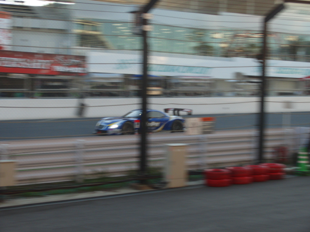 Ein blauer Sportwagen fährt auf einer Rennstrecke entlang, umgeben von einem Zaun, Pfölen, Dröhnen, Verkehrskegeln und anderen Gegenständen. Im Hintergrund sind Gebäude und Schilder mit Text zu sehen, alles leicht unscharf.