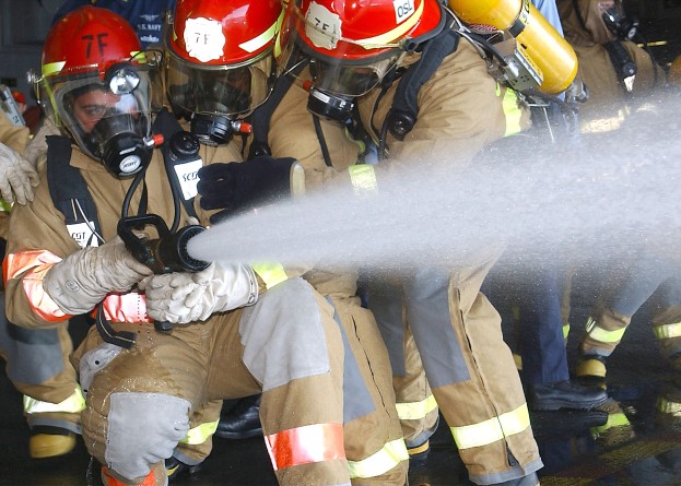 Feuerwehrleute in Helmen, Handschuhen und Gasmasken sprühen Wasser aus einem Schlauch auf den Boden mit einer Wand im Hintergrund.