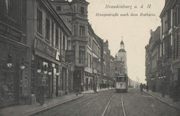 Ein altes Schwarz-Weiß-Foto einer Straßenkreuzung in Brandenburg, Deutschland, mit einer Straßenbahn auf den Schienen, Menschen auf dem Gehweg, Gebäuden mit Fenstern, Schildern, Straßenlaternen und einem bewölkten Himmel.