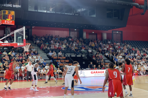 Eine Gruppe von Männern, die Basketball in einer Turnhalle mit einem Netz auf der linken Seite spielen, Zuschauer sitzen im Hintergrund und Stadionlampen darüber.